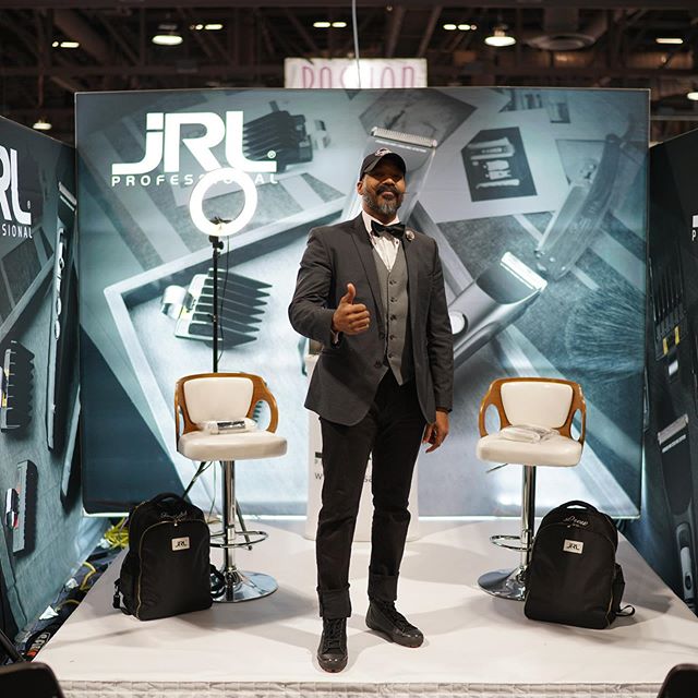 Man in suit and cap giving thumbs up at a professional hair tool booth with two chairs and backpacks.