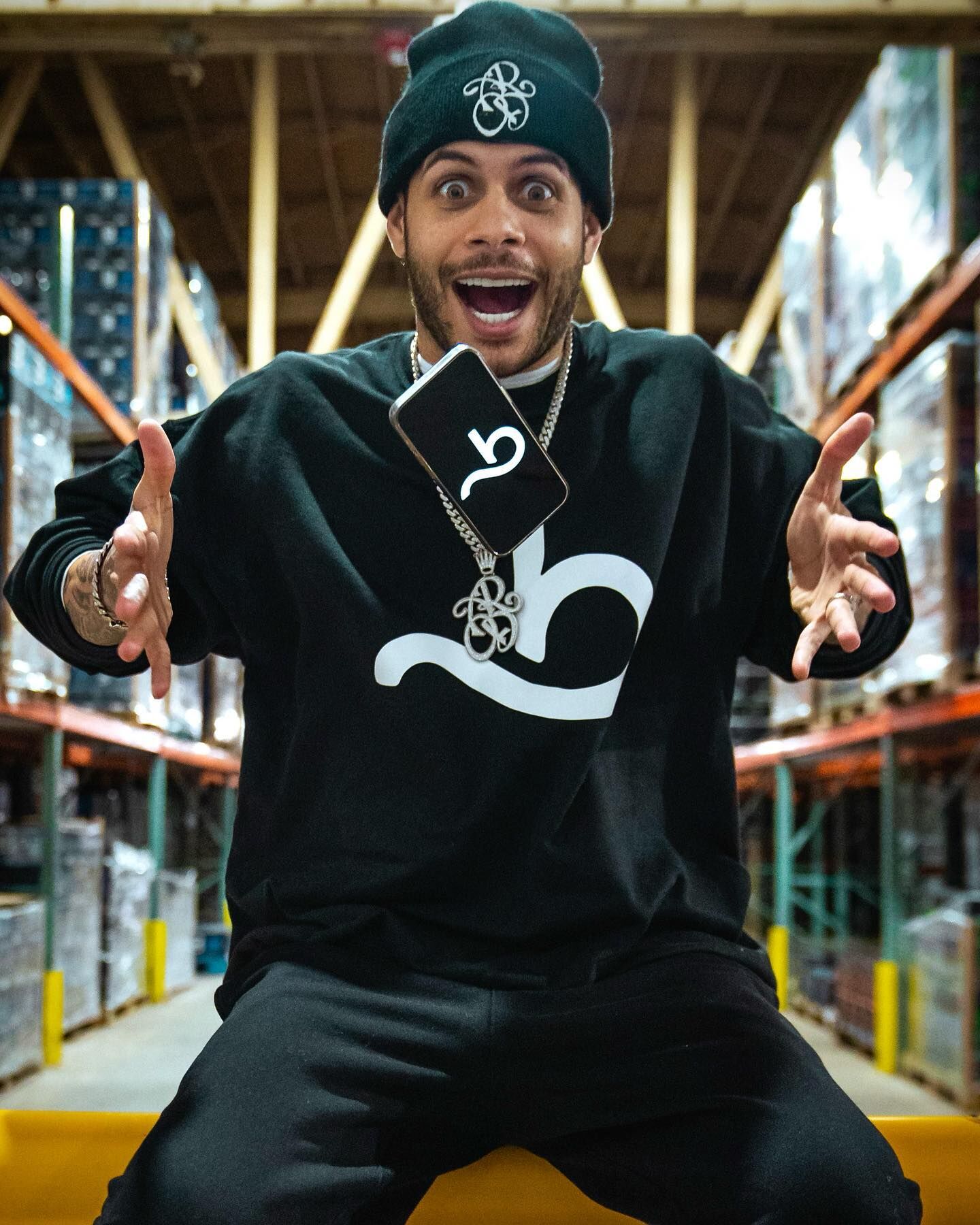 Excited man in black beanie and sweatshirt with a stylized 'b' logo in a warehouse aisle.