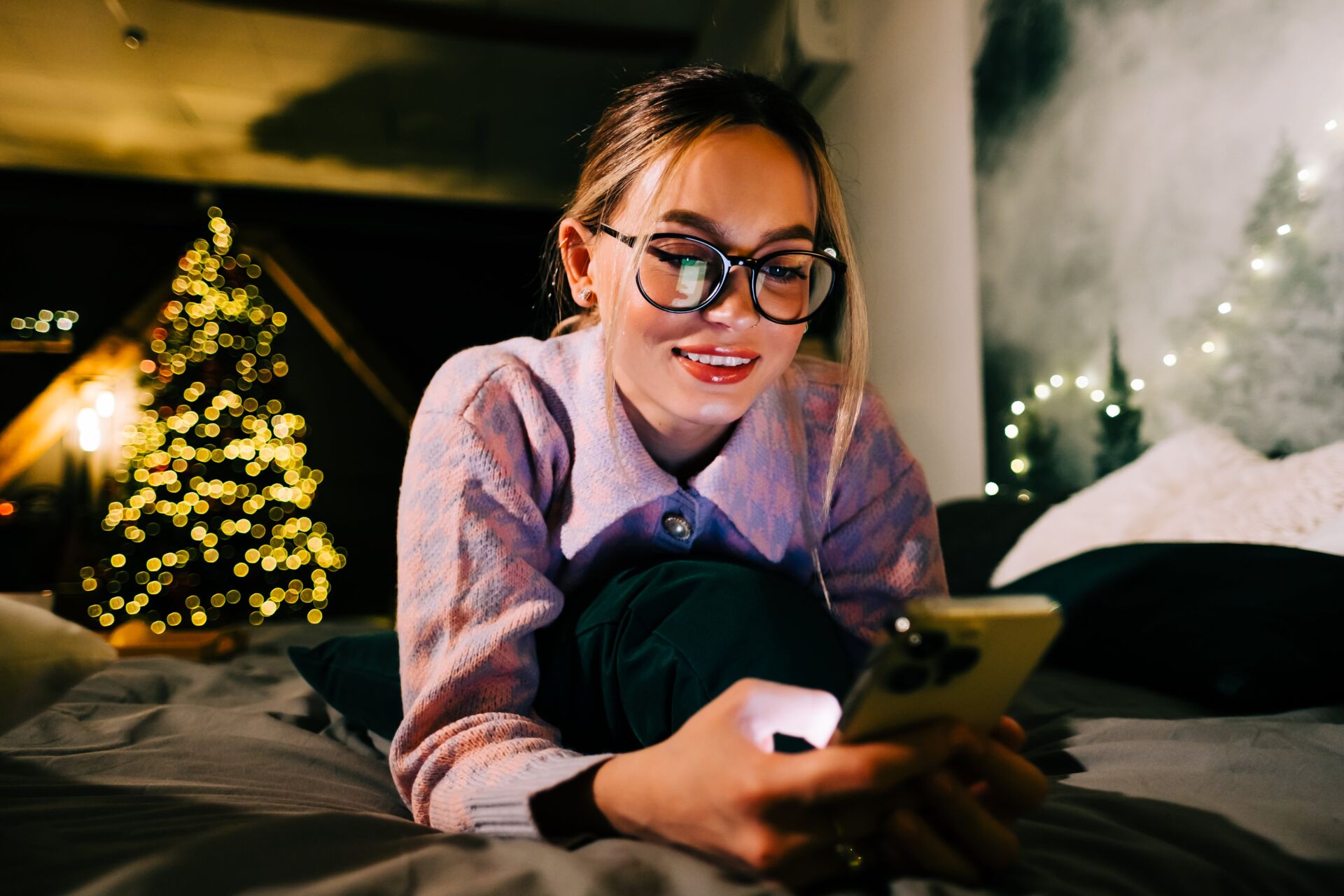 Smiling woman with glasses lying on bed using smartphone with Christmas lights blurred behind her.