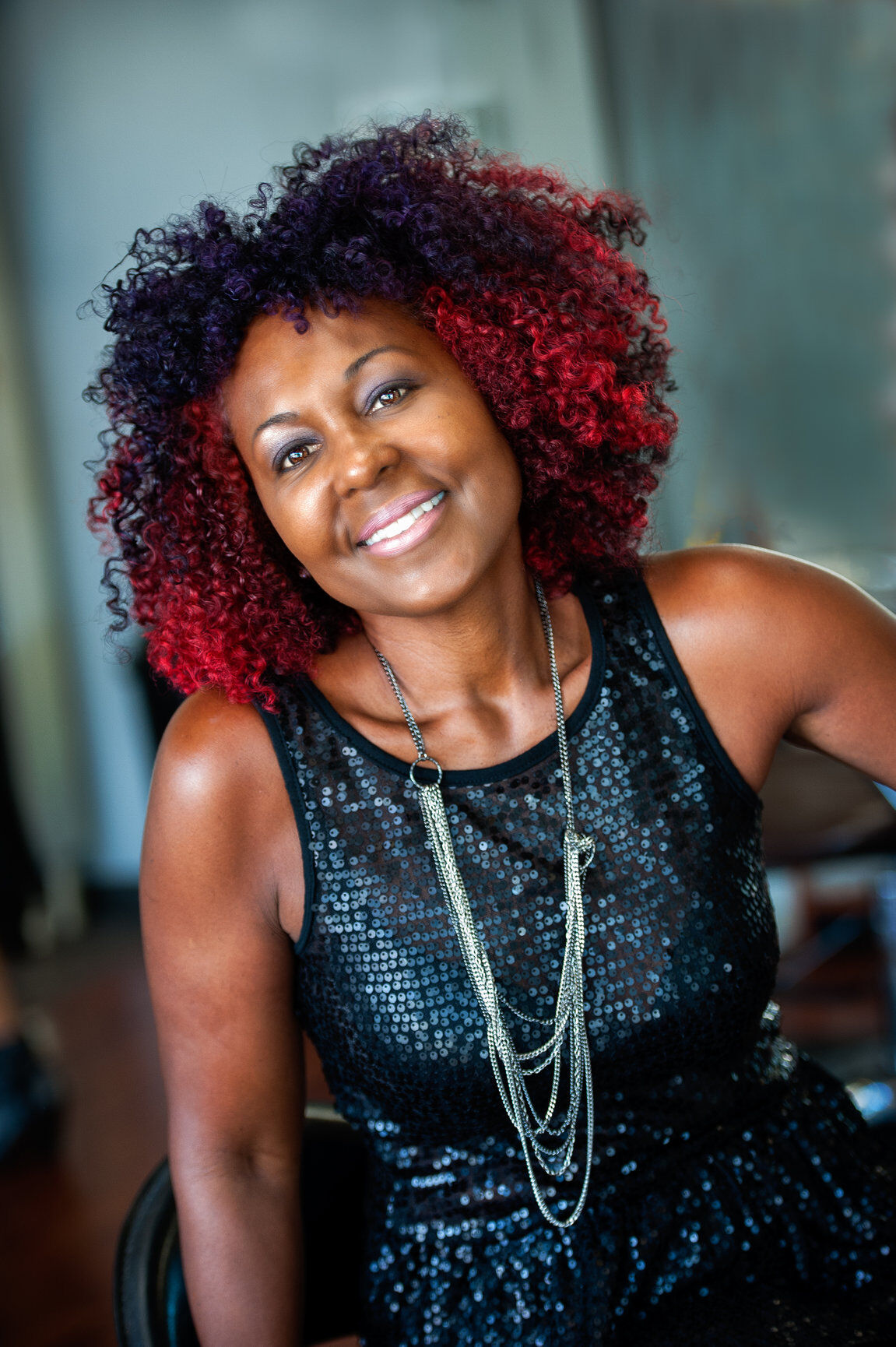 Smiling woman with curly red and black hair wearing a black sequined top and layered silver necklace.