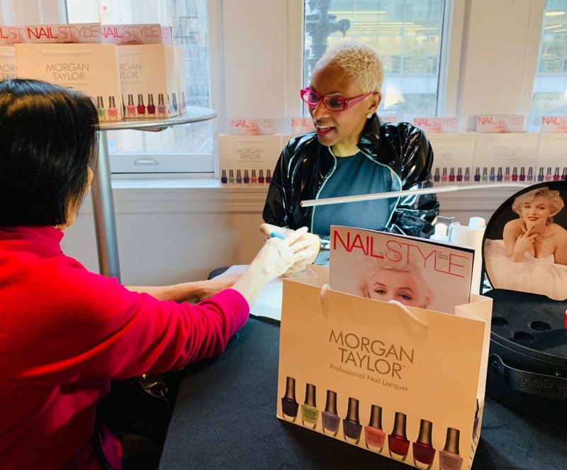 Two women at a nail salon with Morgan Taylor nail polish displayed on the table and windowsill.