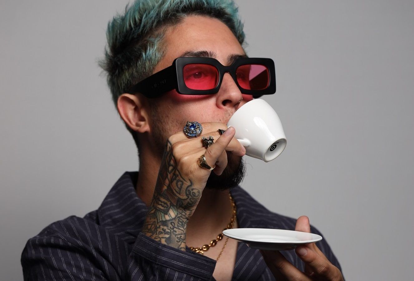 Person with teal hair and tattoos wearing red sunglasses drinking from a white teacup.