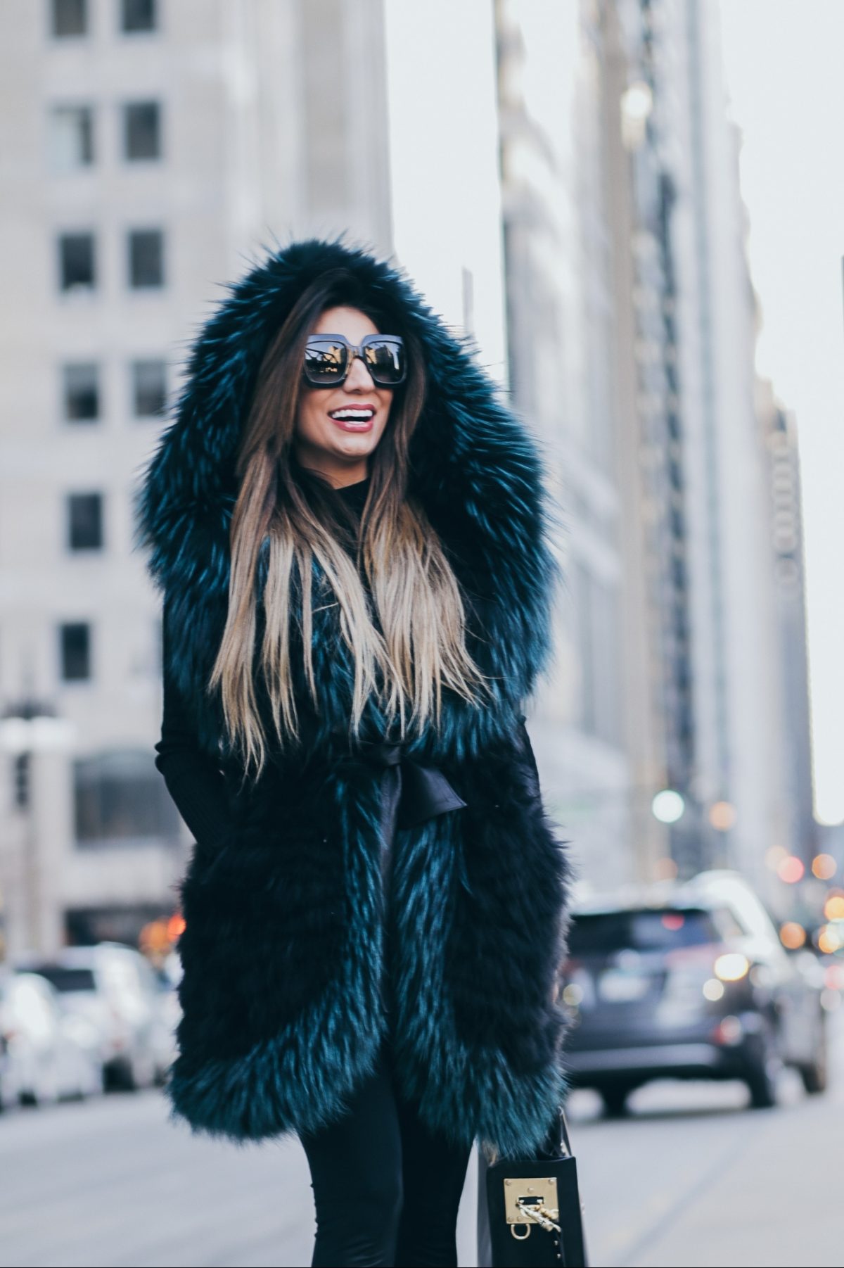 Woman wearing large hooded fur coat and sunglasses, smiling on city street with buildings and cars behind.
