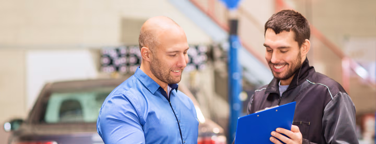 Two men smiling and discussing while looking at a clipboard in an auto repair shop with a car in the background.