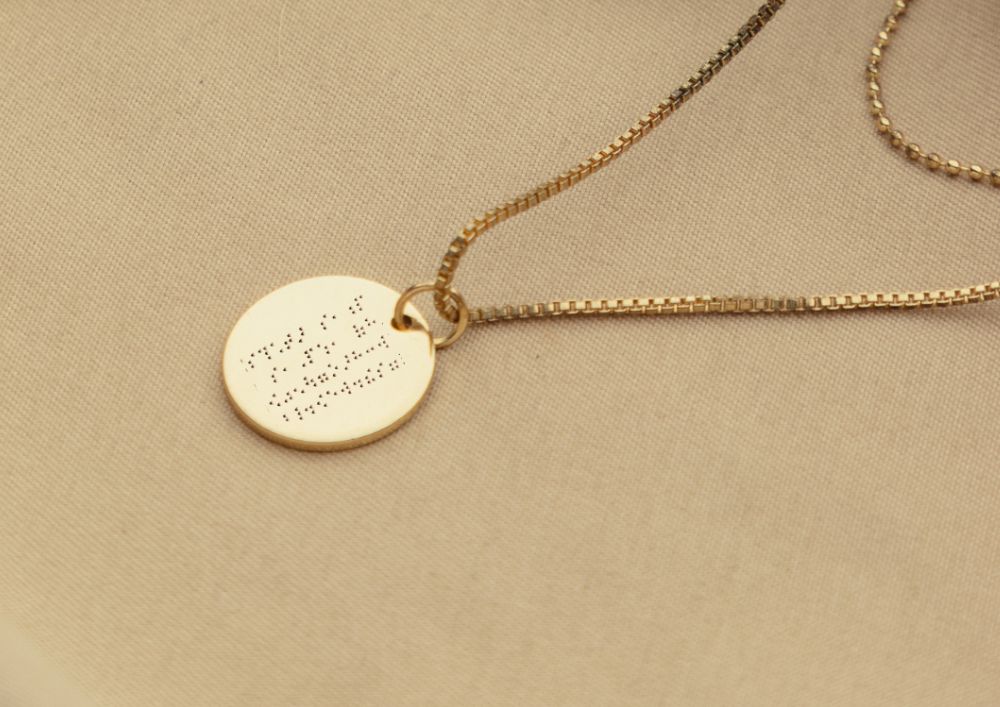 An elegant gold disc necklace engraved with a personal message, including braille text for inclusivity.
