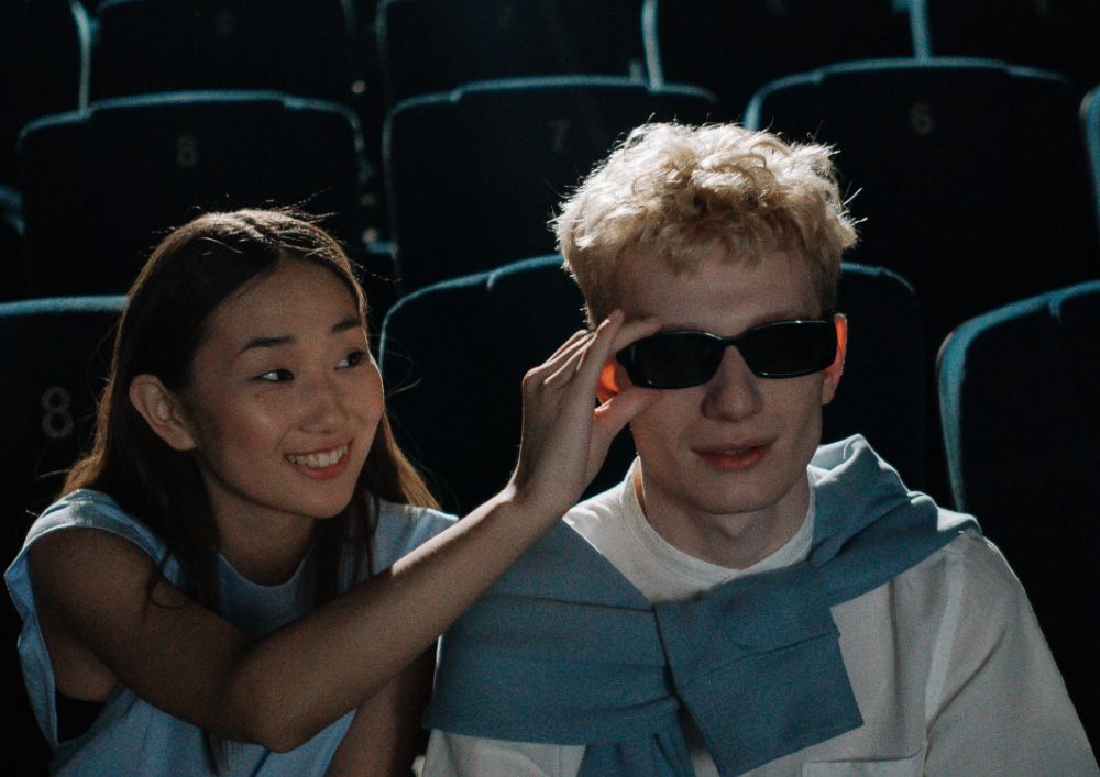 A blind man and a woman wearing sunglasses sit together in a theater, enjoying the show.