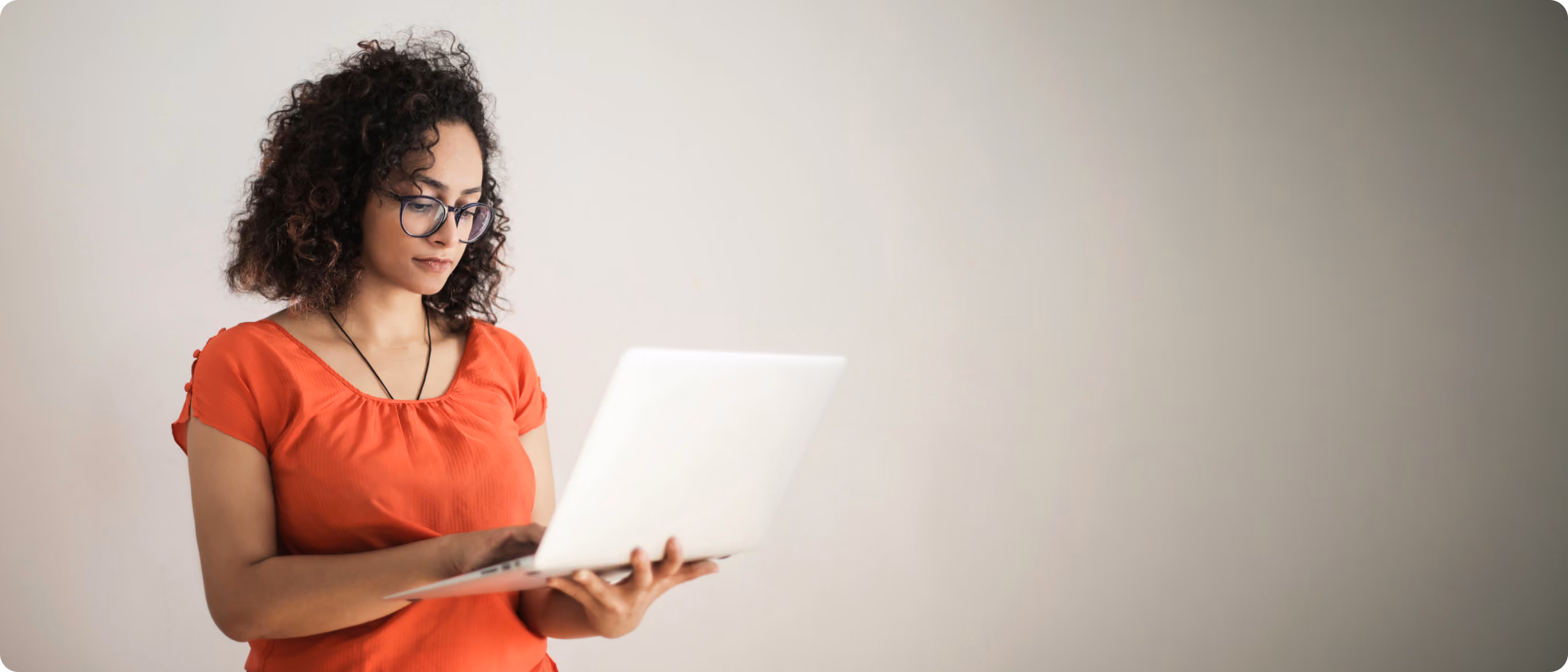 Frau mit lockigem Haar und Brille tippt auf einem Laptop vor neutralem Hintergrund.