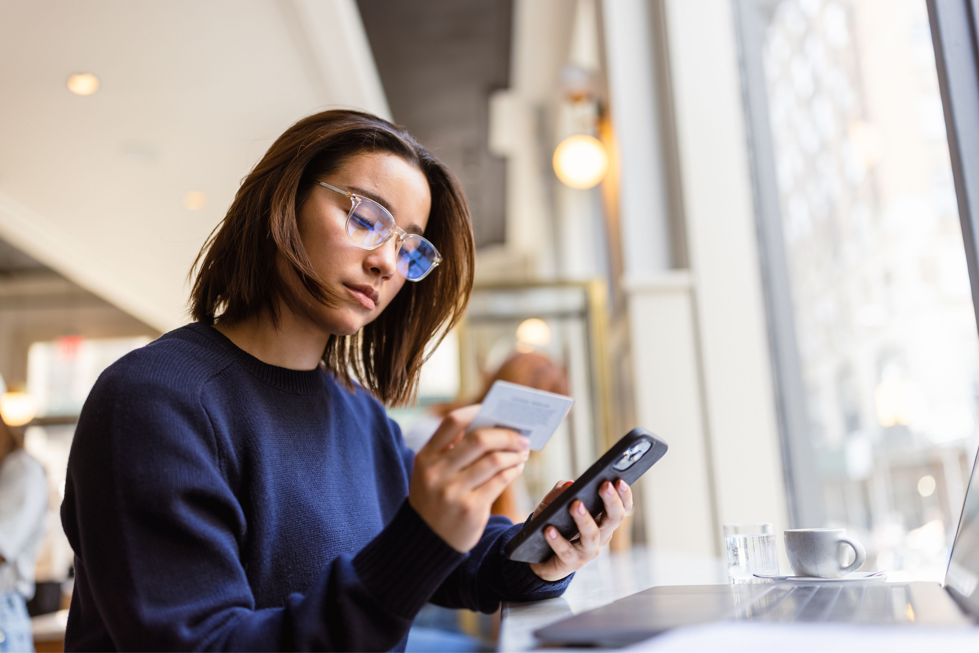 Junge Frau in dunkelblauem Pullover und Brille liest eine Karte, während sie ein Handy hält, an einem Tisch mit Laptop, Tasse und Glas Wasser in einem Café mit großem Fenster.