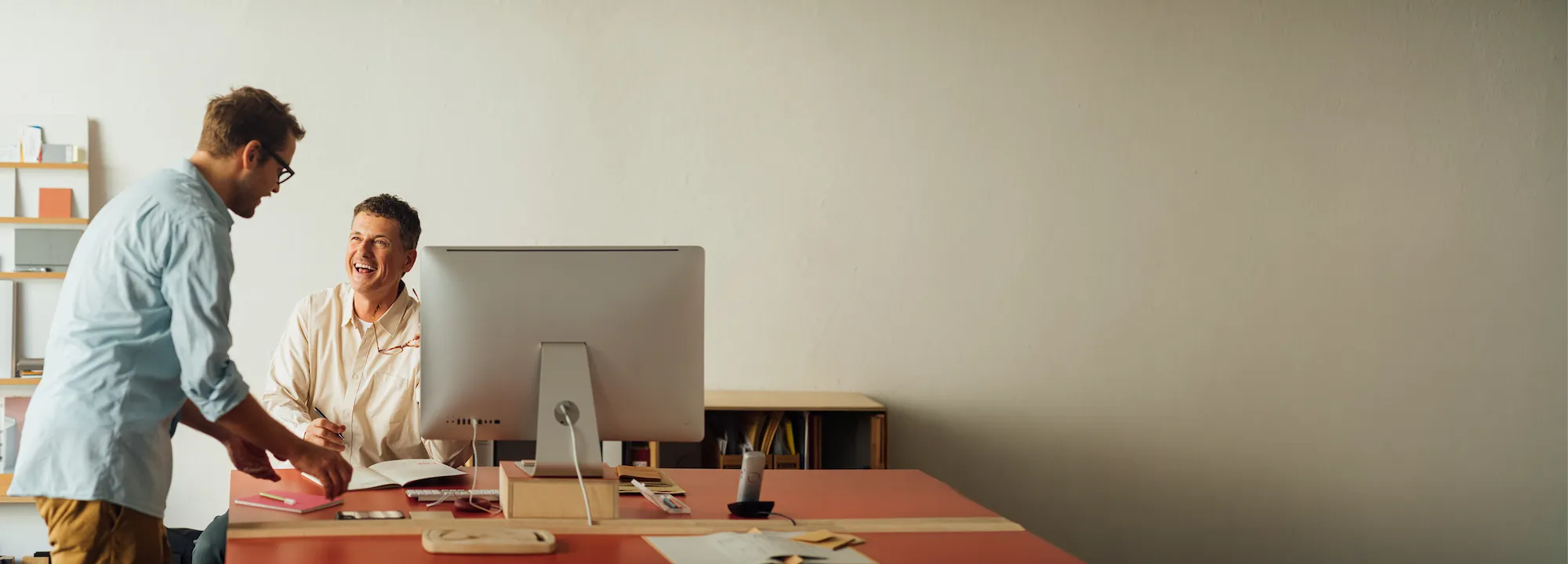 Zwei Männer in einem Büro unterhalten sich, einer sitzt an einem Schreibtisch mit Computer und lächelt, der andere steht und hält ein Notizbuch.