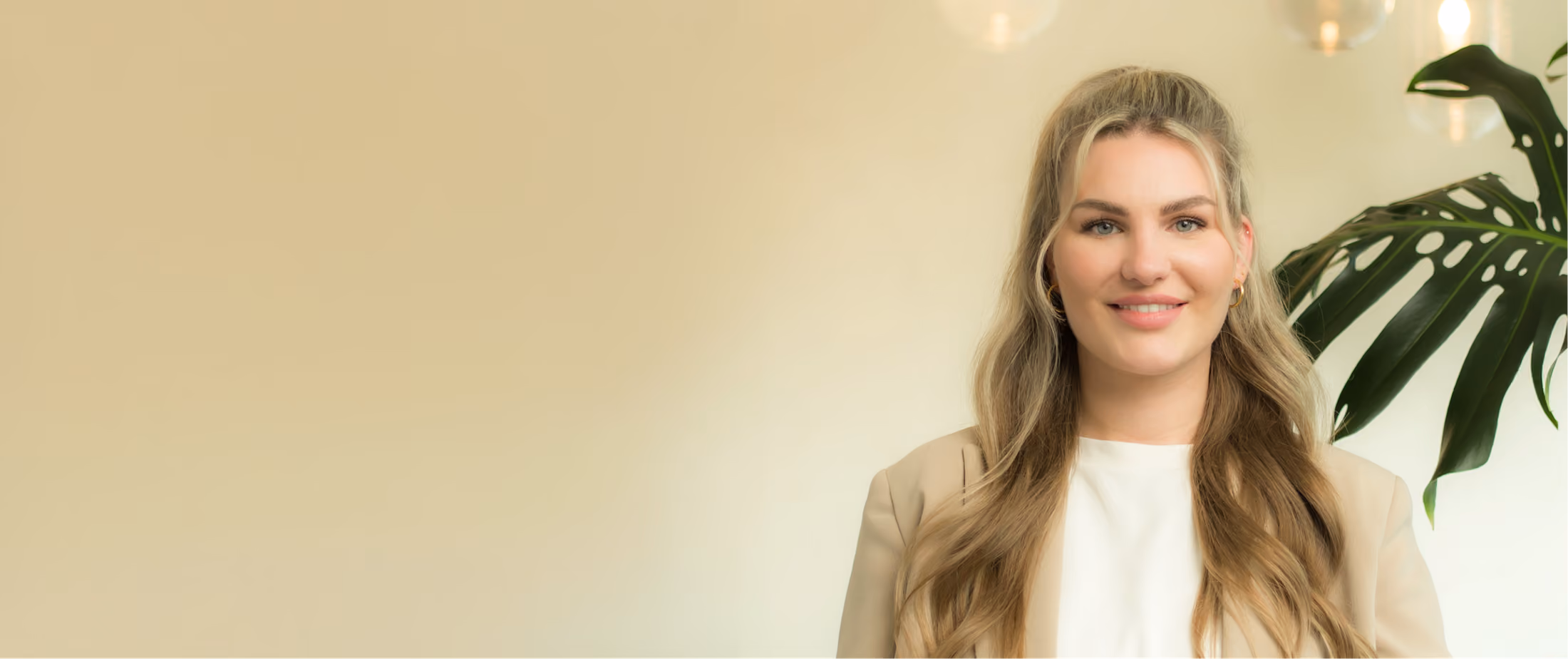 Porträt einer lächelnden jungen Frau mit langen blonden Haaren, die ein helles Oberteil und ein beiges Jackett trägt, vor einem hellen Hintergrund mit einer großen grünen Pflanze rechts.