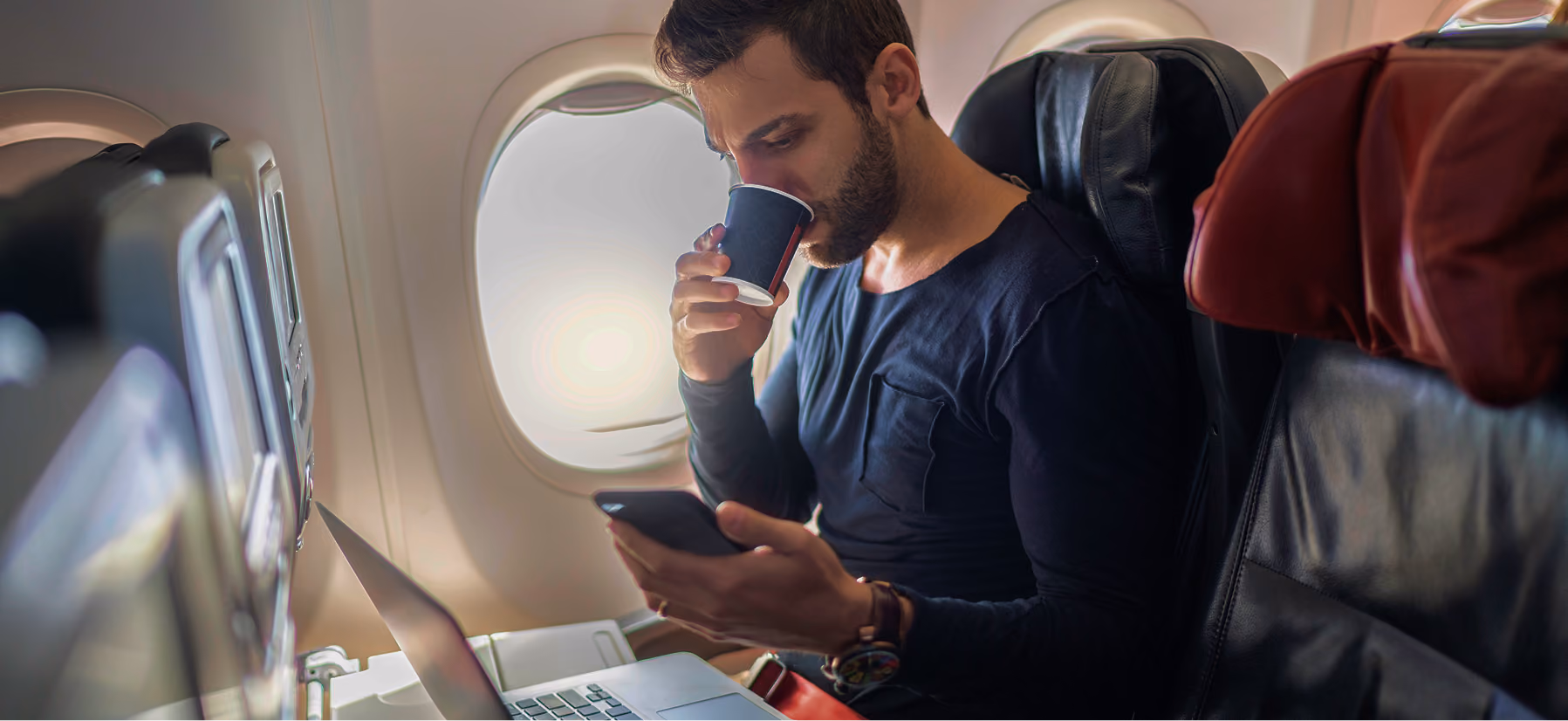 Mann sitzt in einem Flugzeugsitz, trinkt aus einem Pappbecher und sieht auf sein Handy, während ein Laptop vor ihm auf dem Tisch steht.
