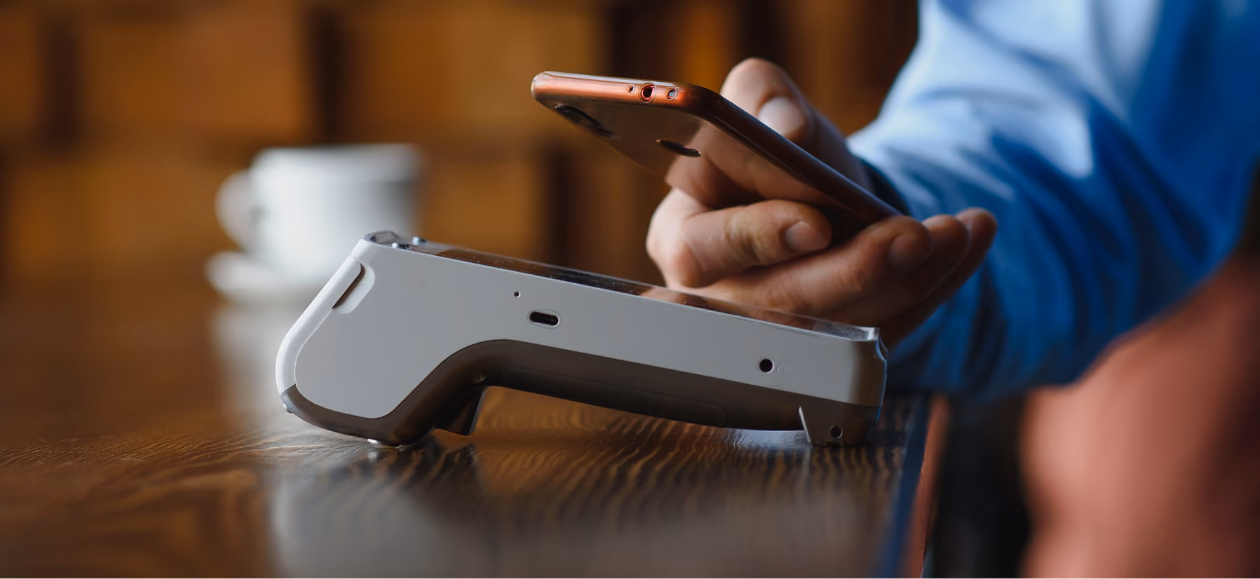 Eine Person hält ein Smartphone nahe an ein kontaktloses Zahlungsmobilgerät auf einem Holztisch, um eine Zahlung durchzuführen.