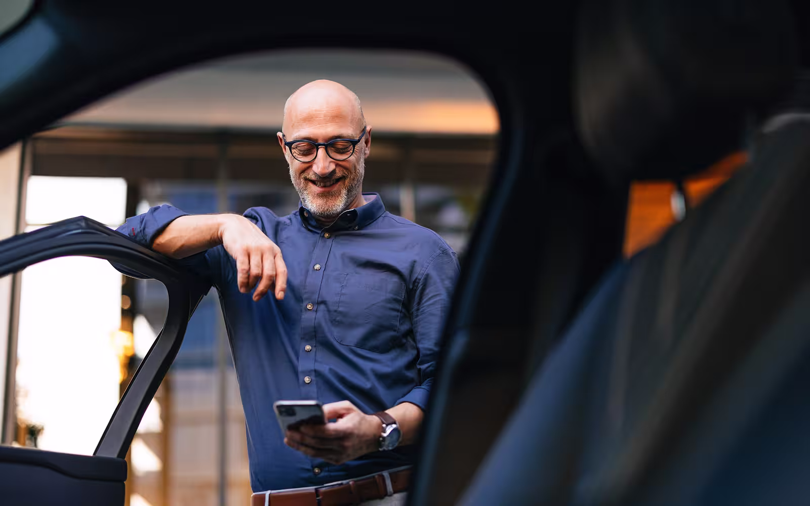 Ein Mann steht an seinem Auto und nutzt lächelnd sein Smartphone.