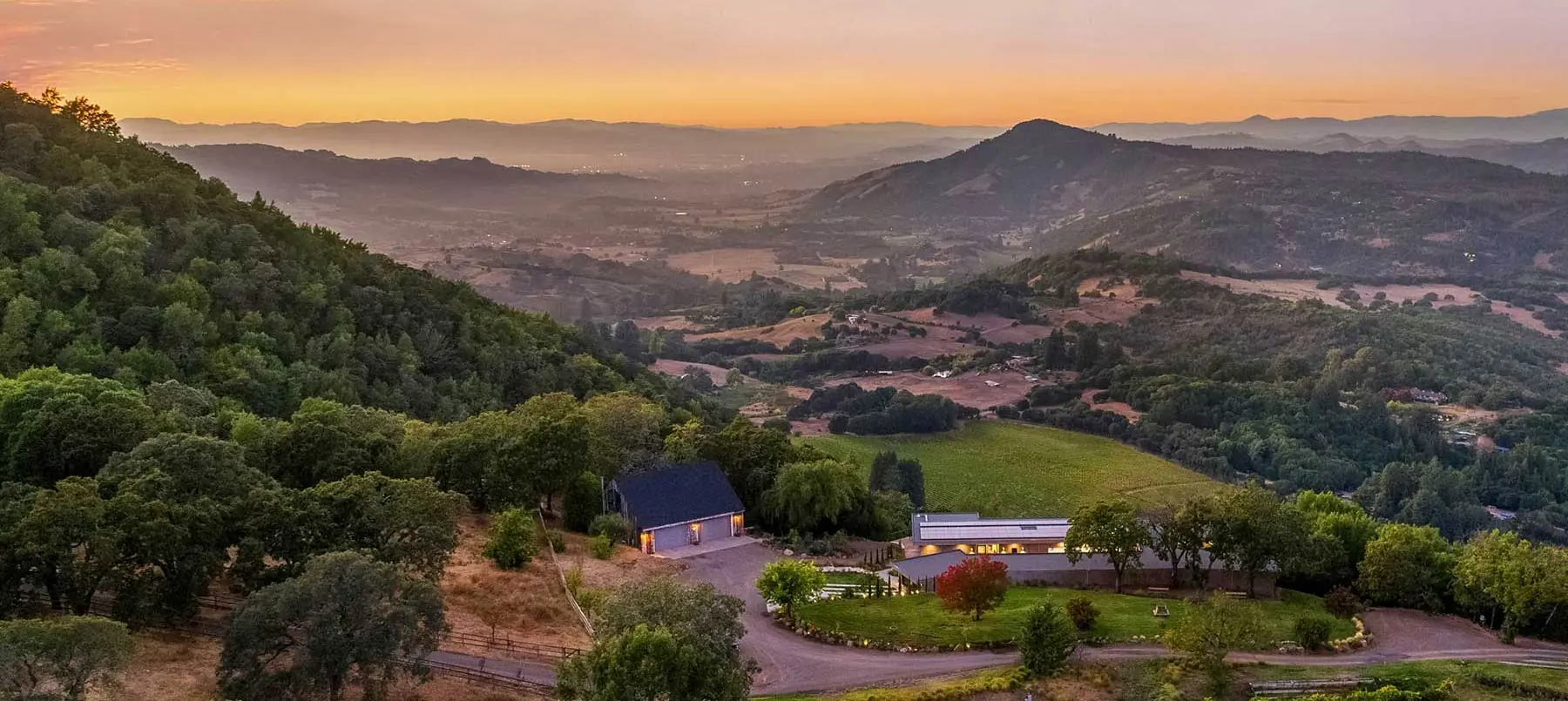Aerial view of a modern hillside estate surrounded by vineyards, rolling hills, and trees at sunset.