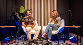 Deux personnes assises sur des fauteuils discutent dans un studio avec des micros sur bras articulés.