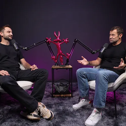 Deux hommes assis sur des fauteuils discutant devant des microphones avec une sculpture rouge au centre.