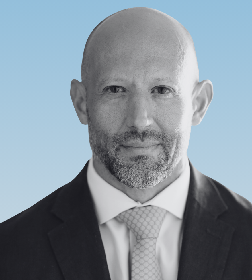 Professional headshot of Christopher Mannino with a close cropped beard and bald head wearing a dark suit white shirt and patterned tie against a light blue background. He faces forward with a steady confident expression.