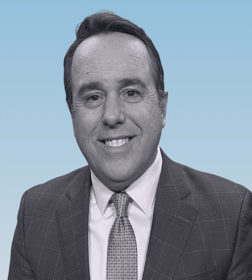 Professional headshot of Ben Bradley smiling with neatly combed hair wearing a windowpane check suit white shirt and textured tie against a light blue background. His friendly expression and direct gaze feel approachable and steady.