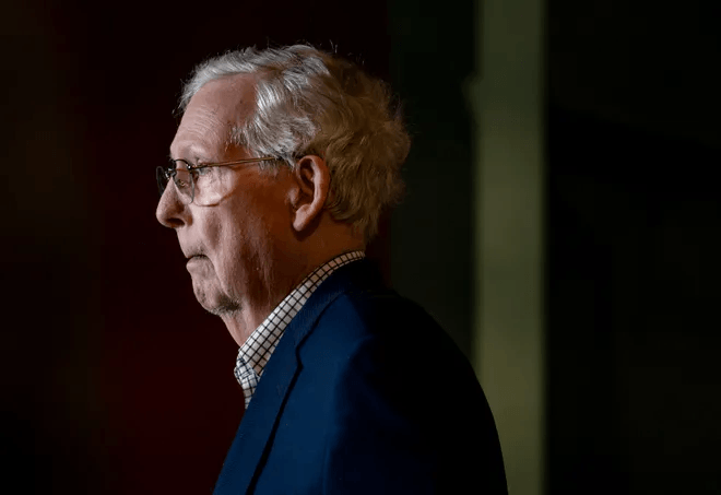 Senator Mitch McConnell in profile wearing glasses and a navy blazer before speaking at a public event.