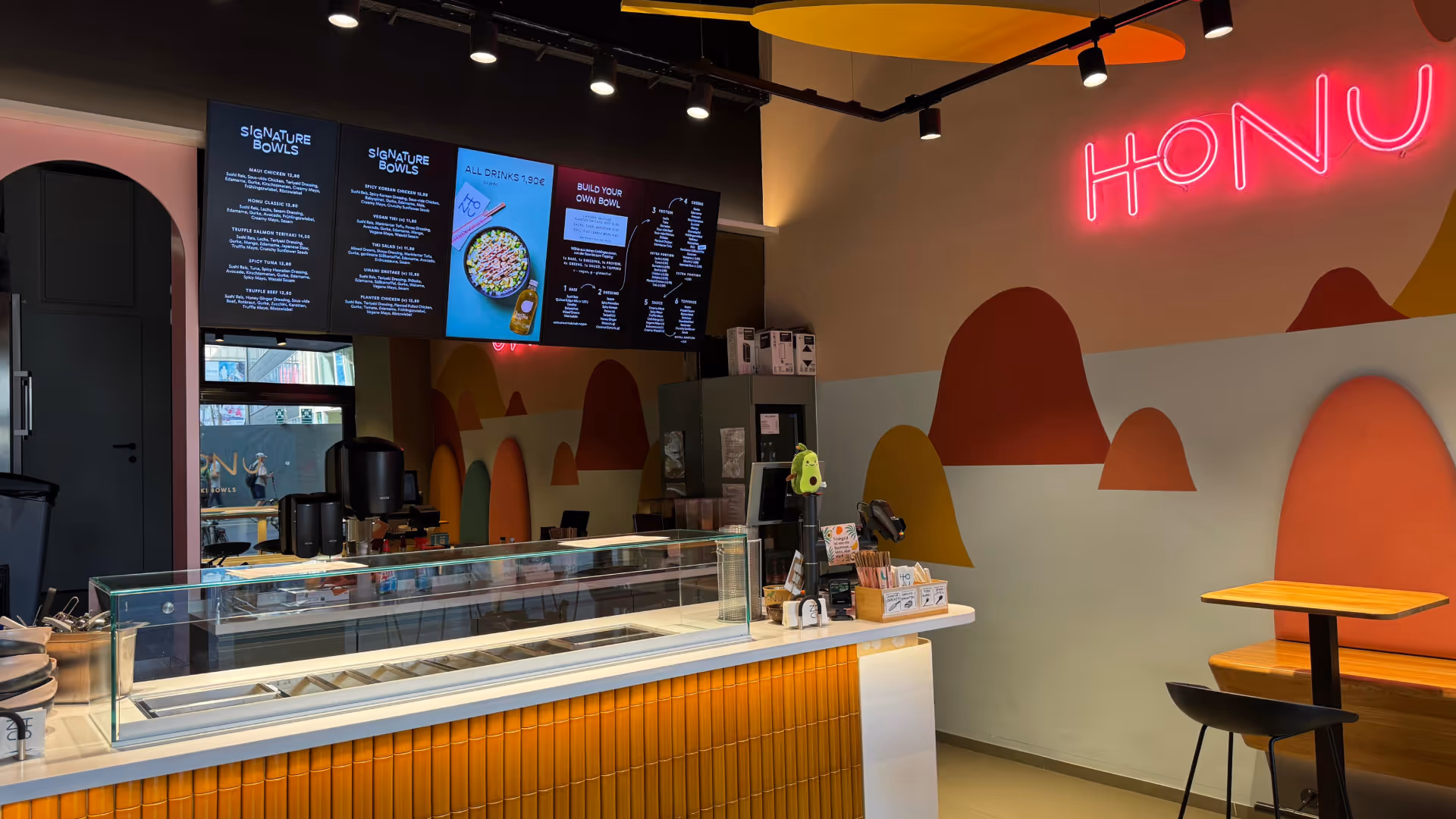Eine moderne Poke-Bar mit farbenfrohem Interieur, Glastheke, digitalen Menübildschirmen und einem neonfarbenen „HONU“-Schild. Das Ambiente ist lebendig und einladend.