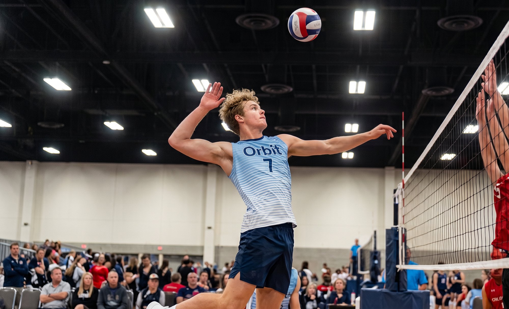 A volleyball player in an Orbit jersey rising to attack the ball at the net, with blockers reaching on the other side