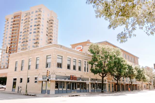 outside of the aiden hotel with a large building in the back with oak trees in the front