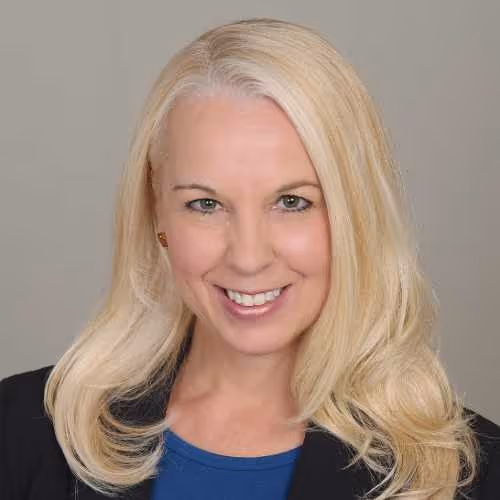 Smiling woman with long blonde hair wearing a black blazer and blue top against a gray background.