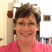 Smiling woman with short dark hair and glasses wearing a pink top in a room with bookshelves and a plaque on the wall.