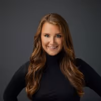 Smiling woman with long wavy brown hair wearing a black turtleneck against a dark gray background.