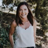Smiling woman with dark hair wearing a white sleeveless top standing outdoors with greenery in the background.