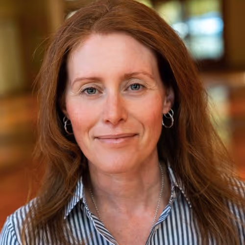 Portrait of a woman with long reddish-brown hair, wearing hoop earrings and a striped shirt, smiling softly indoors.