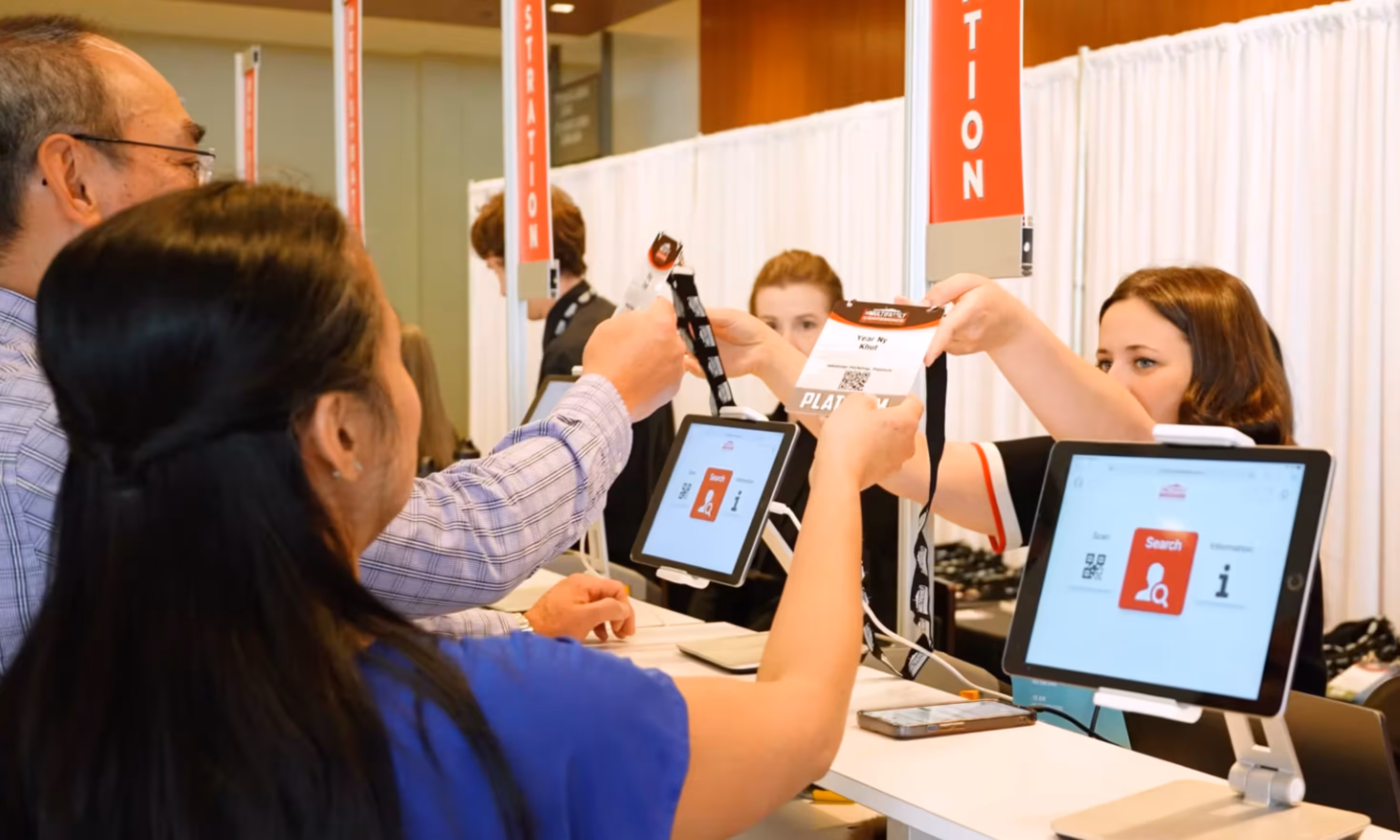 Event attendees receiving badges from staff at a registration desk with tablets and red registration signs.