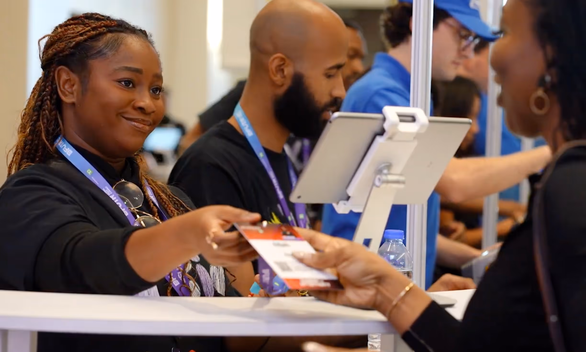 Smiling woman handing an event badge to another person at a registration desk with staff and tablets in the background.