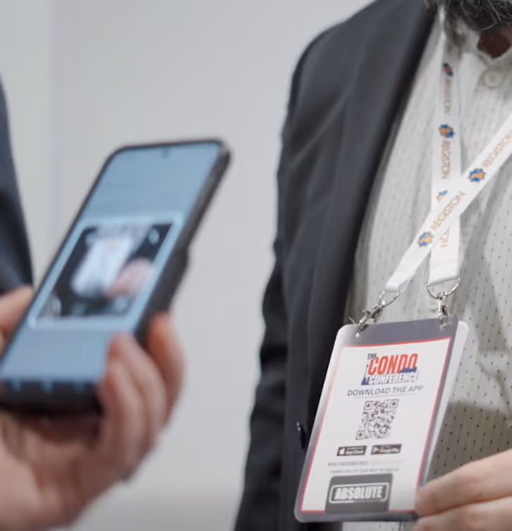 Close-up of a person wearing a conference badge for The Condo Conference with QR code and lanyard, another person holds a smartphone nearby.