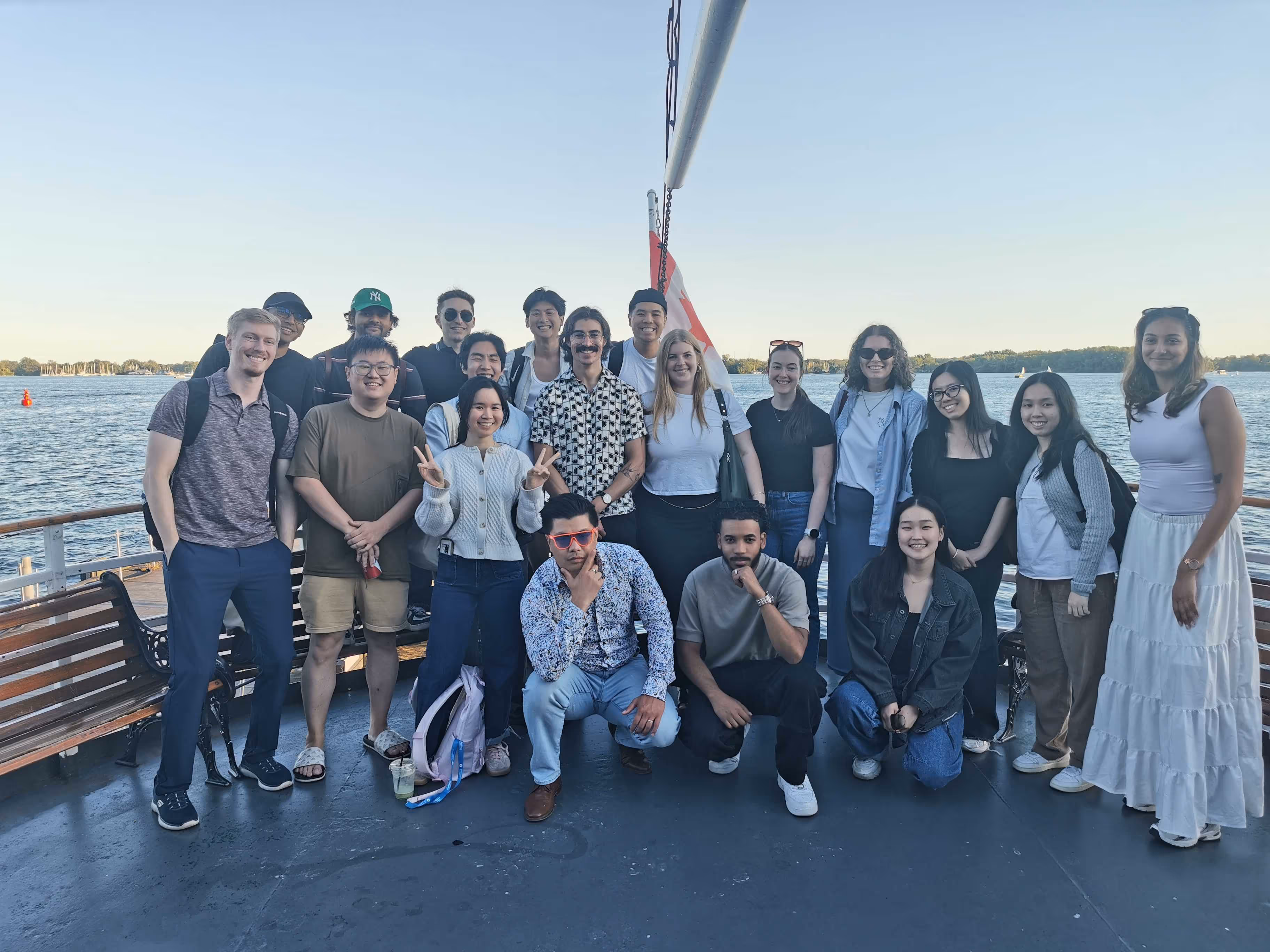 Group of diverse young adults posing and smiling together on a boat deck with water and a clear sky in the background.