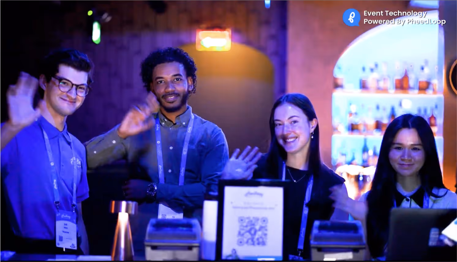 Four smiling event staff behind a lit counter waving to the camera in a dimly lit venue with blue and purple lighting.