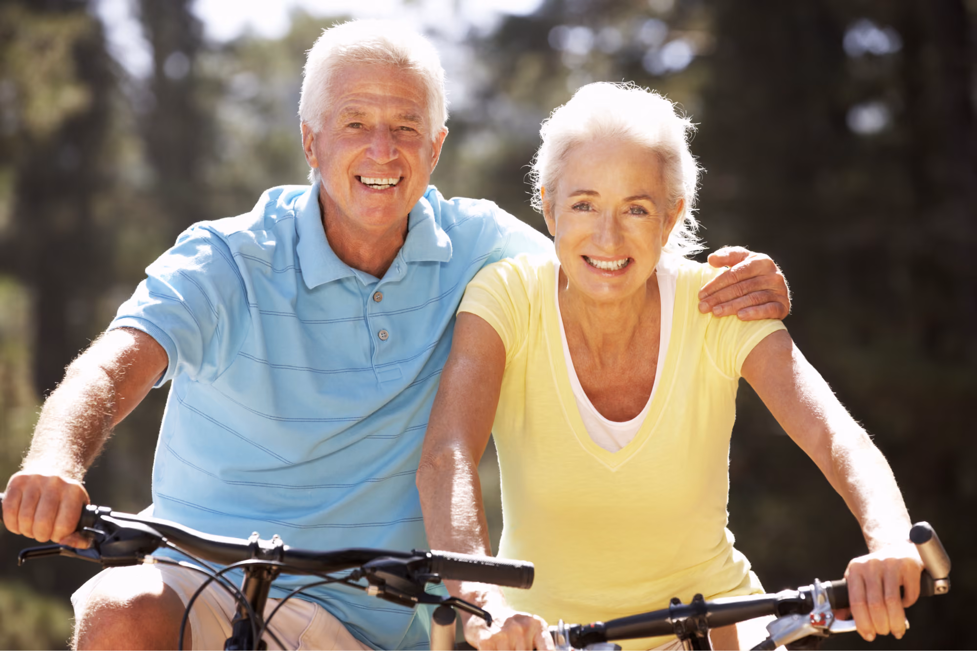 A joyful older couple, both smiling broadly, ride bicycles outdoors while embracing on a sunny day, representing an active and healthy lifestyle.