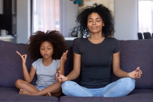 A mother and young daughter meditating together on a couch, representing strong family mental health and wellness supported by self-care strategies.