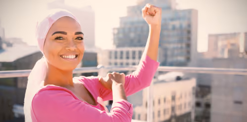 A smiling woman wearing a pink headscarf and pink ribbon flexing her arm, symbolizing strength and positive patient outcomes in oncology rehabilitation.