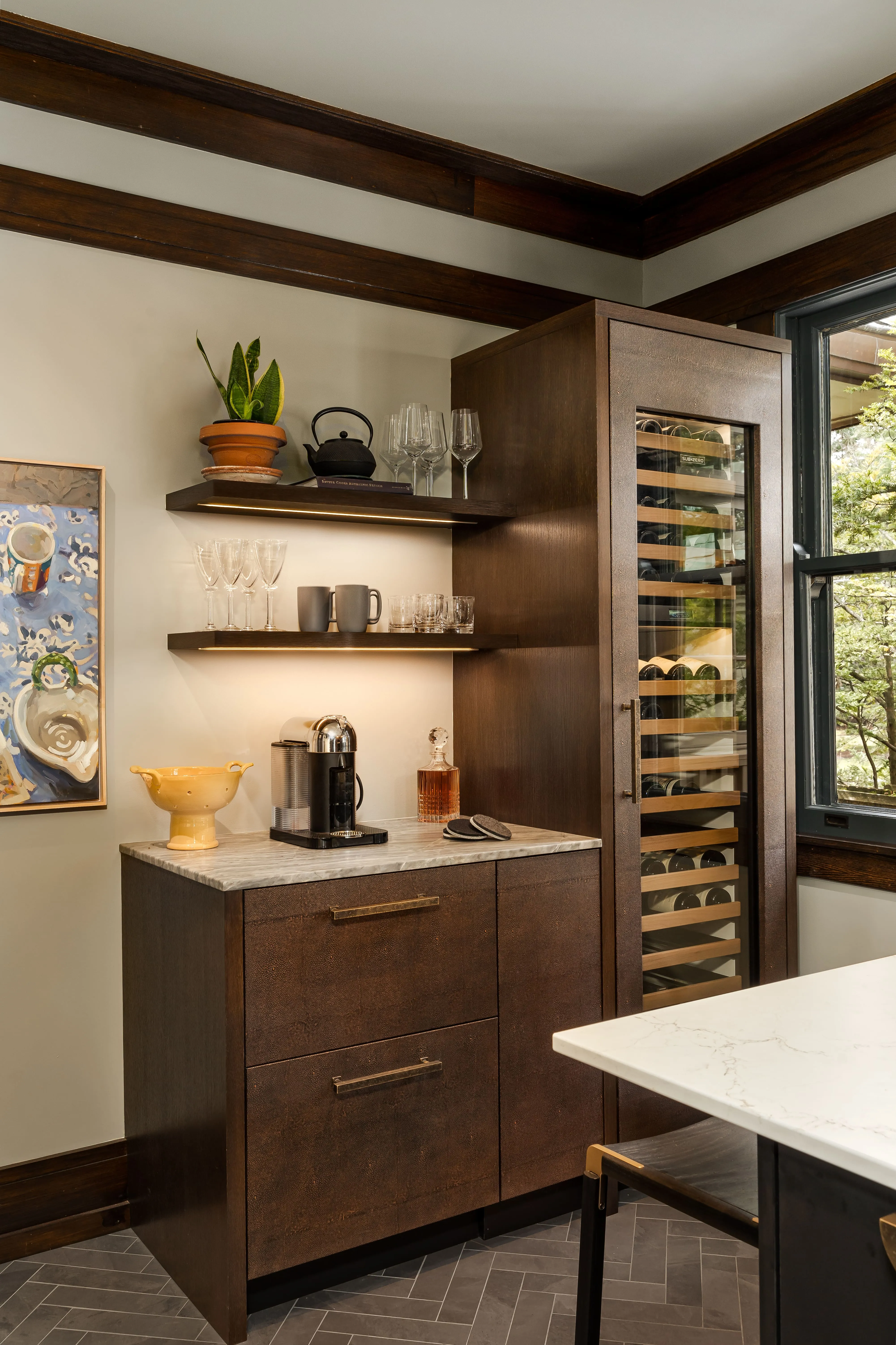 Modern kitchen corner with dark wood cabinets, marble countertop, wine fridge, coffee machine, glassware, and a potted plant on floating shelves.