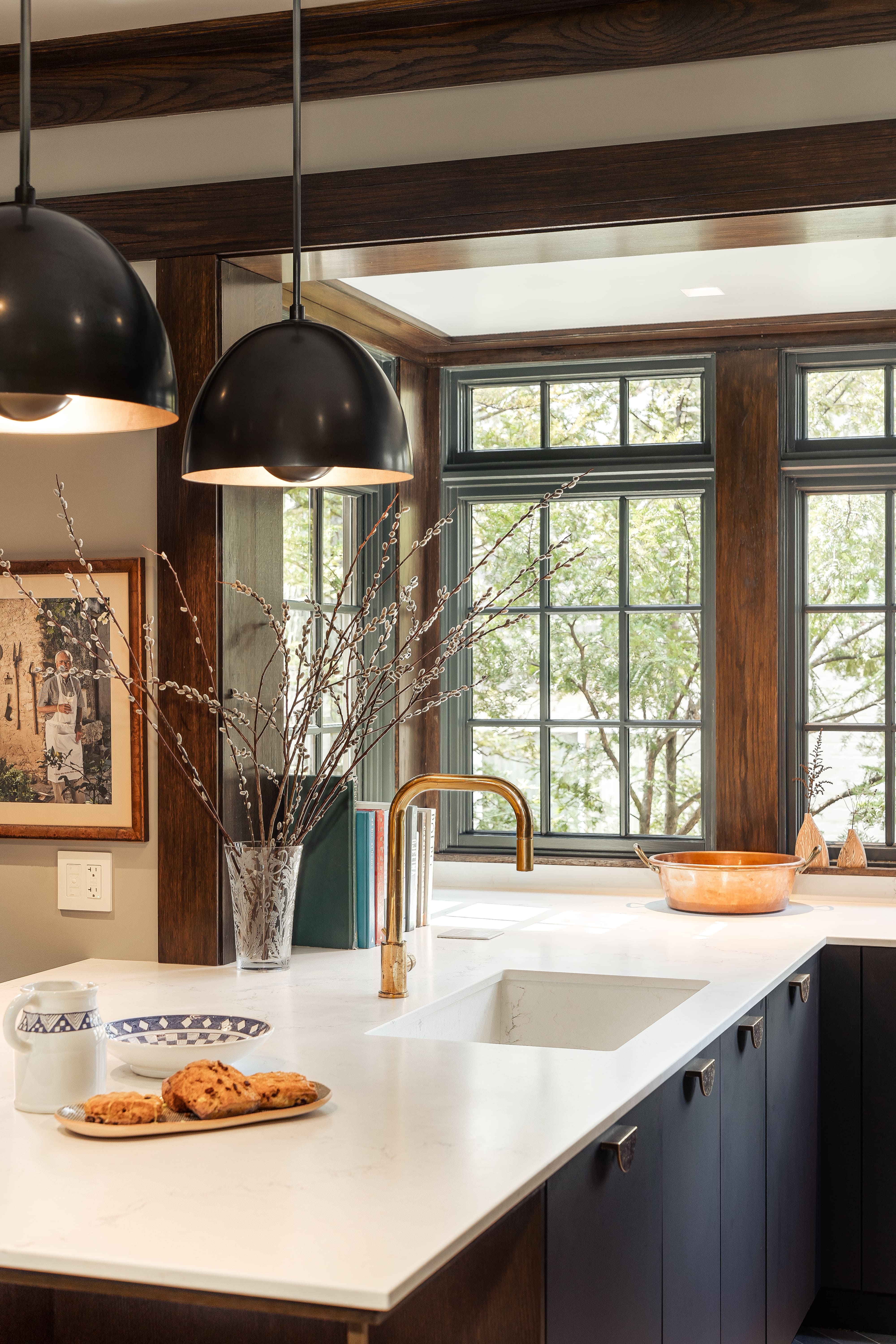 Modern kitchen with dark cabinetry, white marble countertop, brass faucet, large window, hanging black pendant lights, vase with branches, plate of pastries, and copper pot.