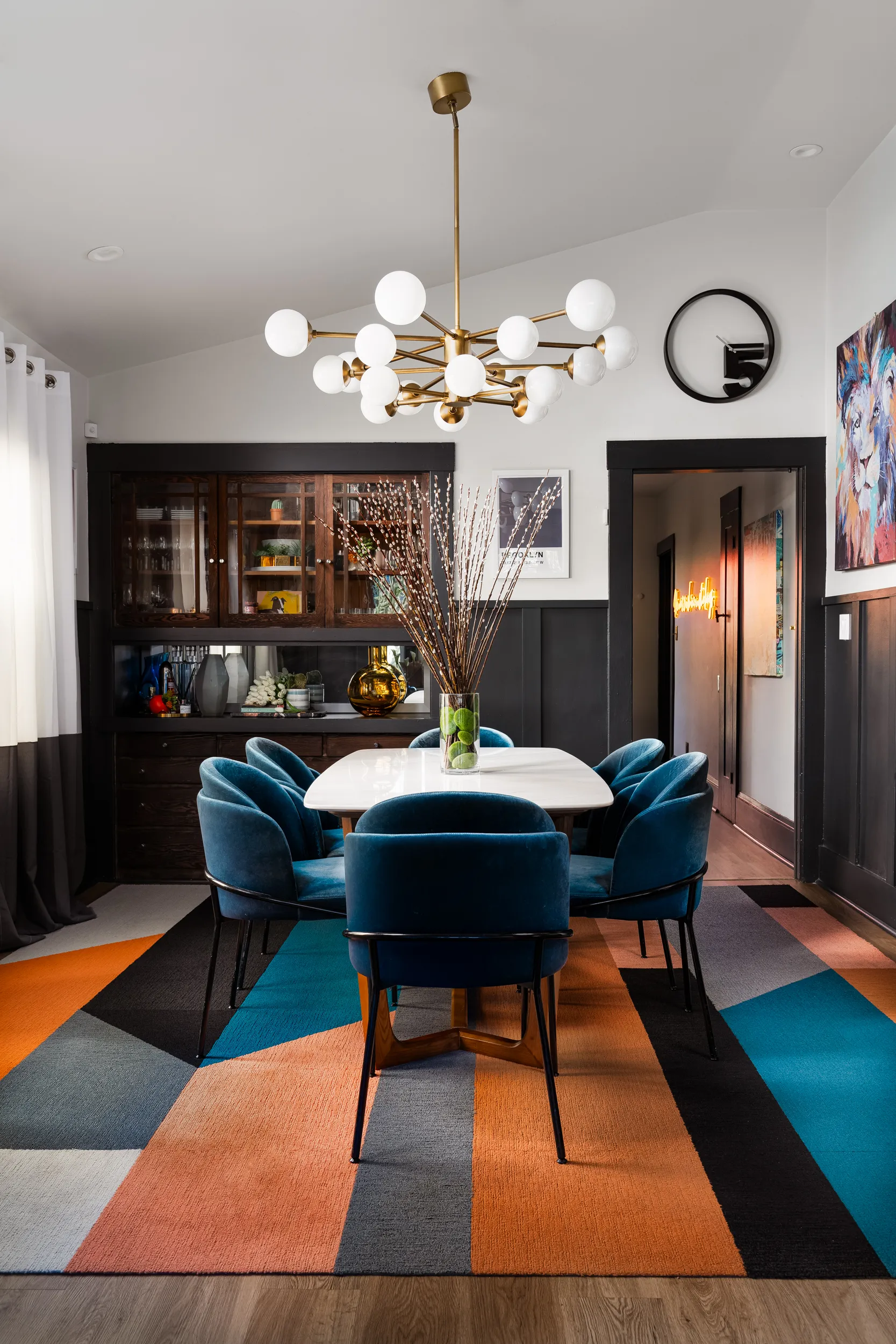 Dining room with a white table, six blue velvet chairs, geometric multicolor rug, a large modern chandelier, and a vase with tall branches in the center.