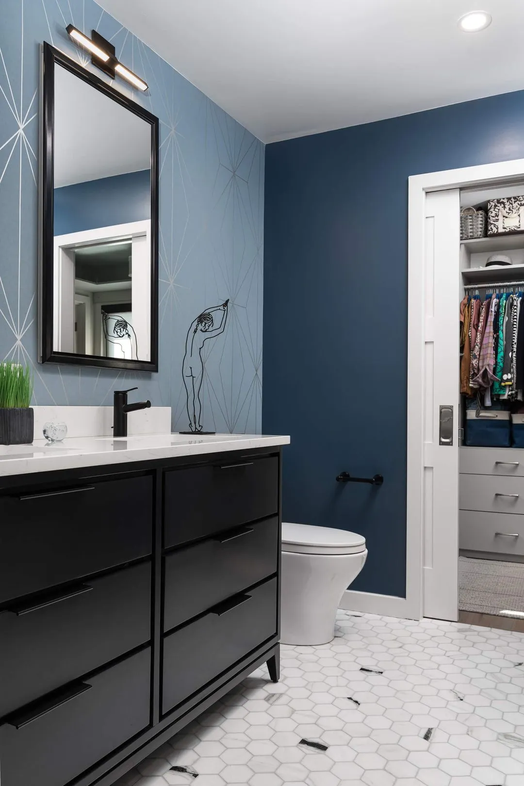 Modern bathroom with black vanity, white countertop, hexagon tile floor, blue walls, wall-mounted mirror, and open closet with clothes.