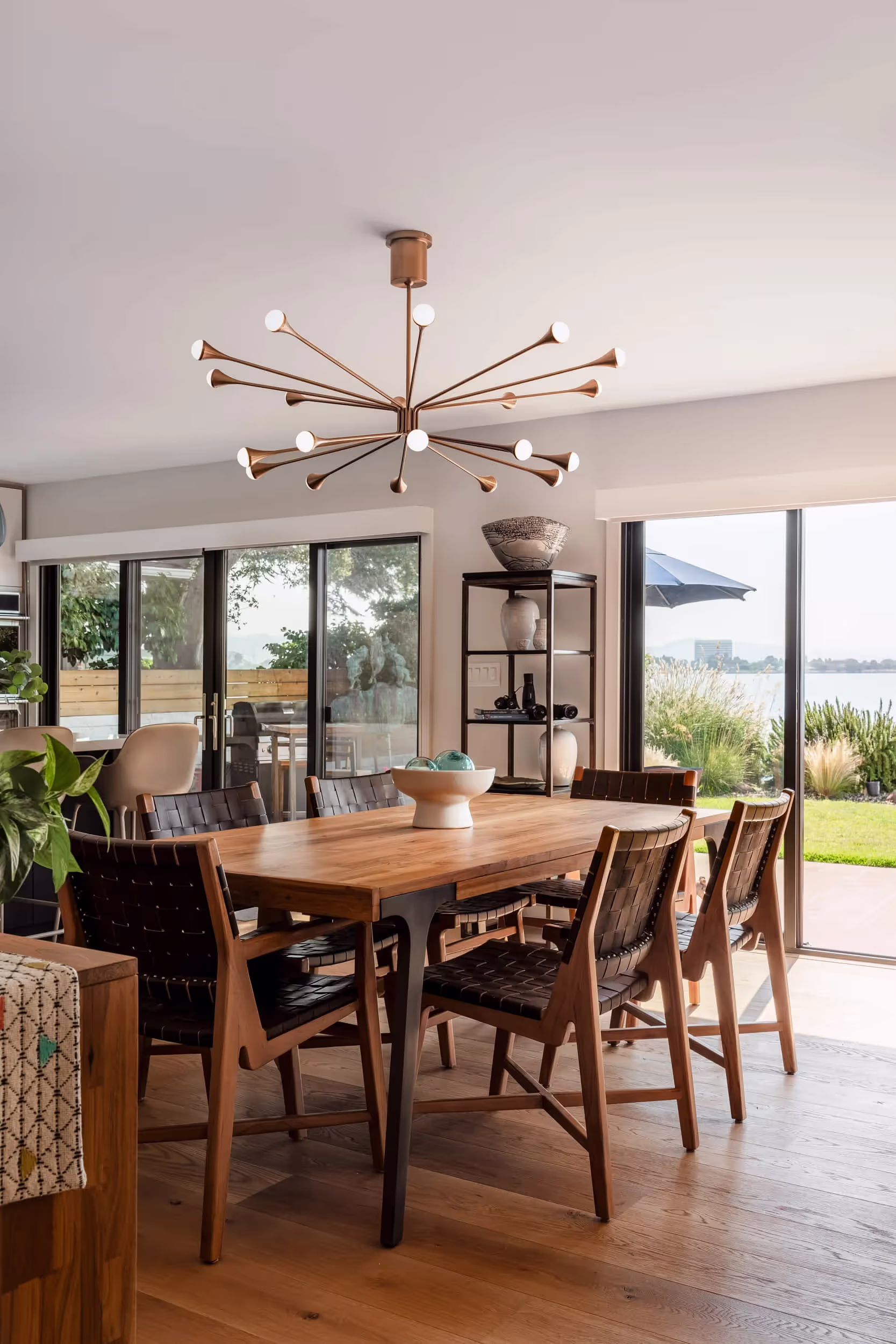 Modern dining area with a wooden table, six leather woven chairs, unique chandelier, and sliding glass doors overlooking a green outdoor space.