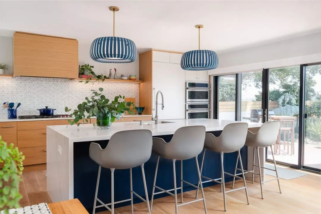 Modern kitchen with a large white marble island, four grey bar stools, blue pendant lights, wooden cabinets, and sliding glass doors opening to an outdoor area.