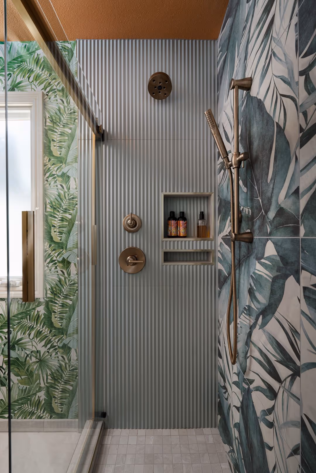 Modern shower with ribbed light blue wall tiles, tropical leaf patterned tiles, bronze shower fixtures, and built-in shelves with bottles.