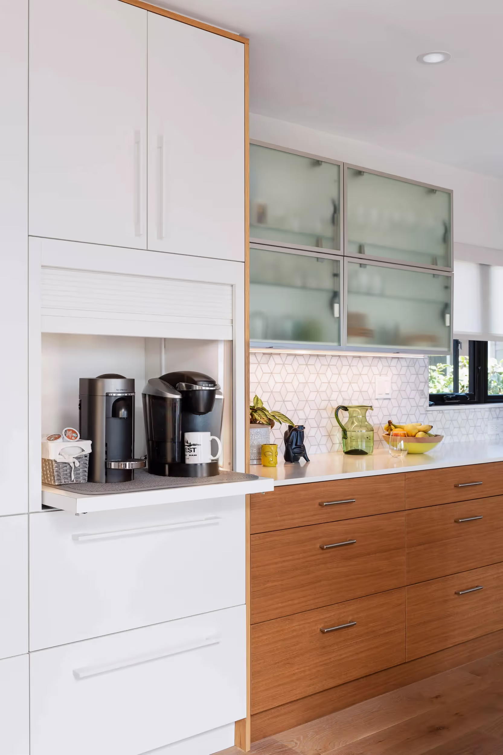 Modern kitchen with white and wood cabinets, featuring a built-in pull-out shelf holding two coffee machines and a white mug.