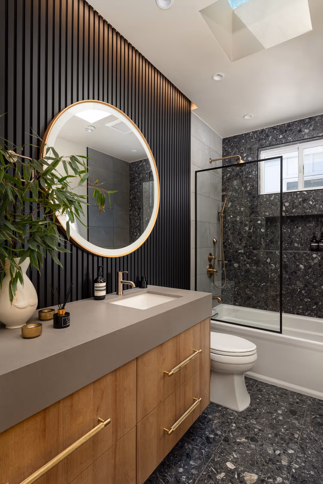 Modern bathroom with wooden vanity, round mirror, black slatted wall, and black and gray tiled shower and floor with a skylight above.