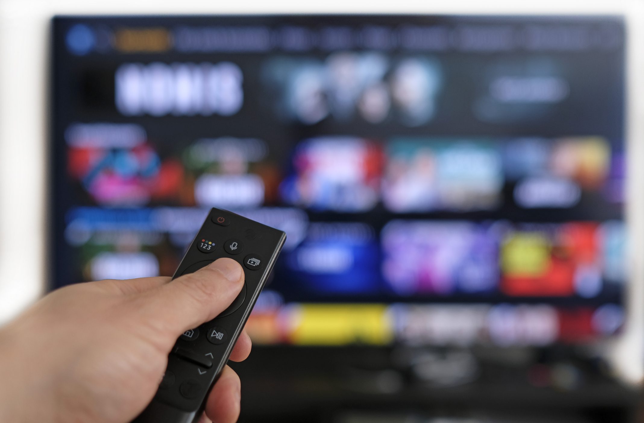 Hand holding a black tv remote and pointing it directly at a flat screen television monitor that has multiple media applications open on the front screen