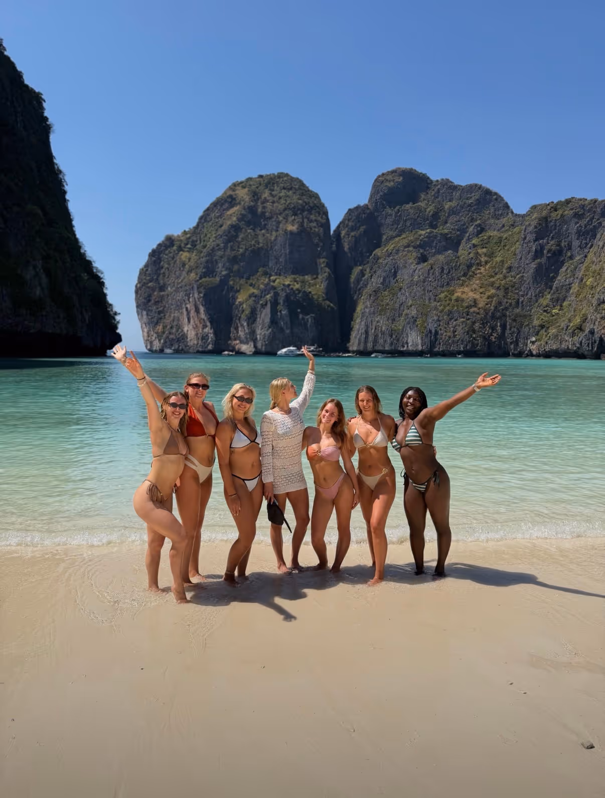 Zes vrouwen in bikini's poseren blij op een zandstrand met turquoise water en rotsachtige eilanden op een zonnige dag.